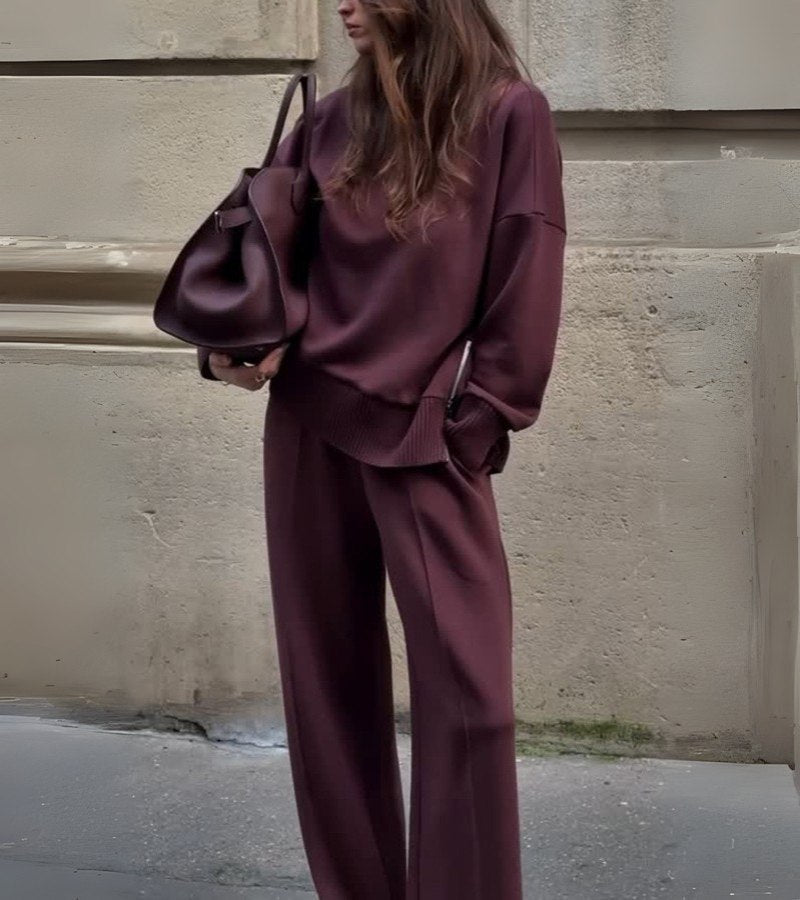 Woman wearing a soft, burgundy two-piece set with a large bag, standing against an urban concrete wall.