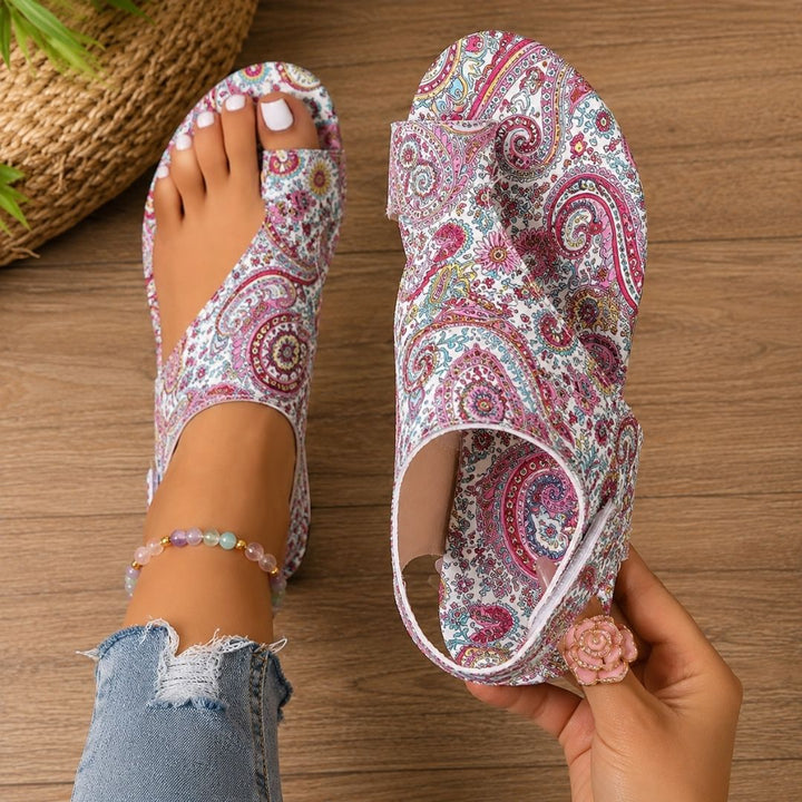 Pink paisley-patterned orthopedic sandals on feet with anklet, denim jeans, wooden floor background.