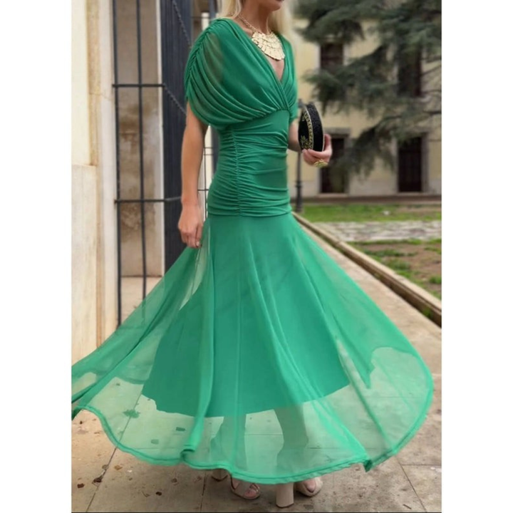 Woman wearing a green mesh dress and statement necklace, holding a black clutch, standing near a railing with trees in the background.