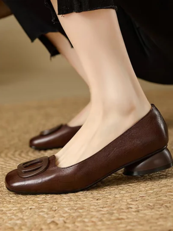 Brown leather flats with prominent buckle design, worn with black pants, on woven mat.