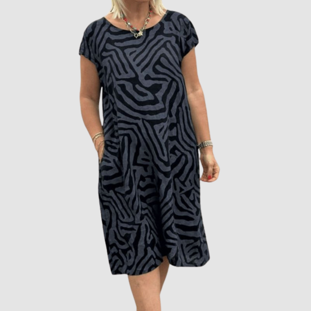 Individual in a dark-toned, vintage-inspired dress with striking abstract pattern, holding pockets, plain backdrop.