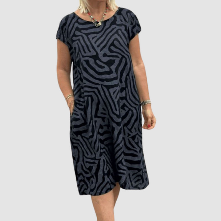 Individual in a dark-toned, vintage-inspired dress with striking abstract pattern, holding pockets, plain backdrop.