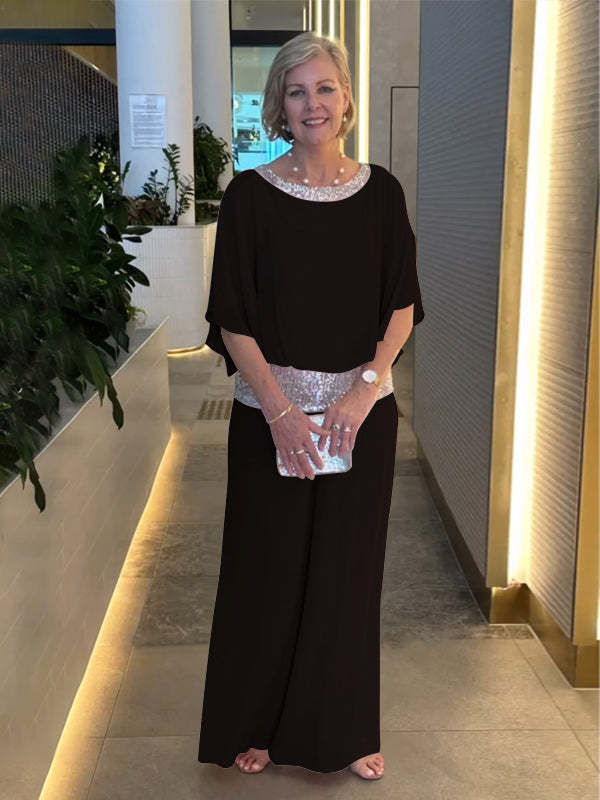 Woman in a black elegant set with shimmering belt and neckline, holding a silver clutch. Indoor setting with plants.