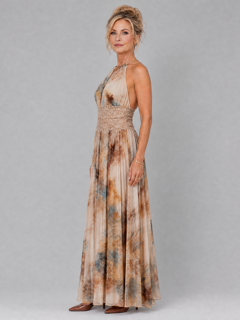 Side view of a woman in a flowing halter neck maxi dress with lace waist, earthy hues, on a grey background.