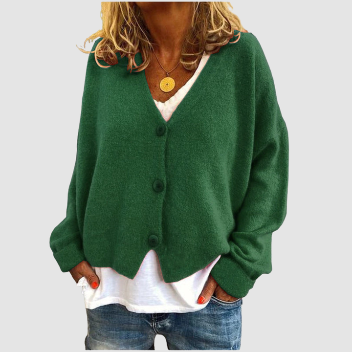 Woman wearing a green knit cardigan over a white shirt and blue jeans, set against a plain gray background.