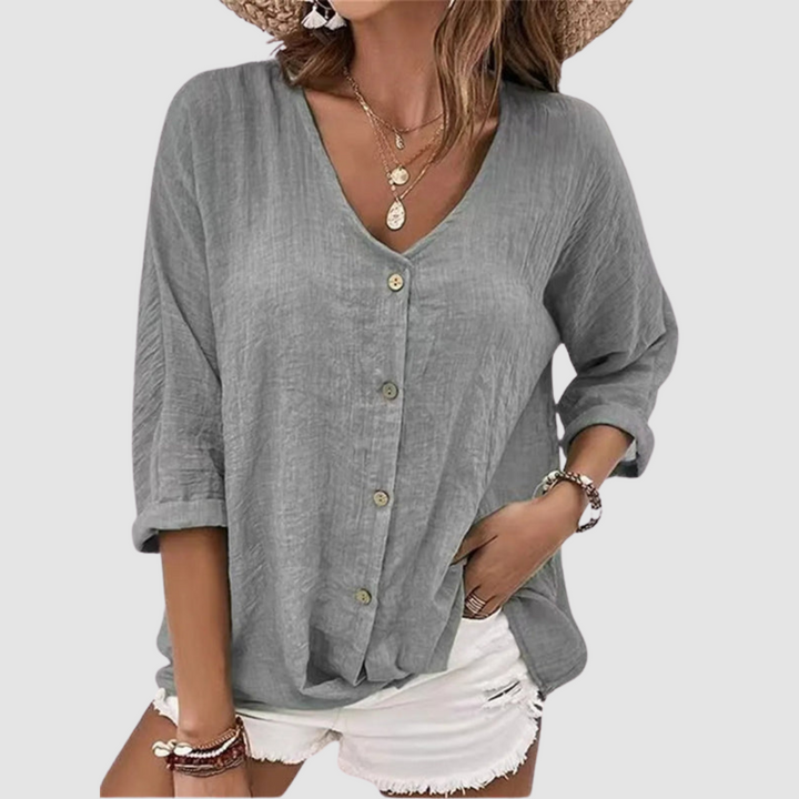 Woman wearing a grey V-neck blouse with buttons, accessorized with necklaces and bracelets, paired with white shorts against a plain background.