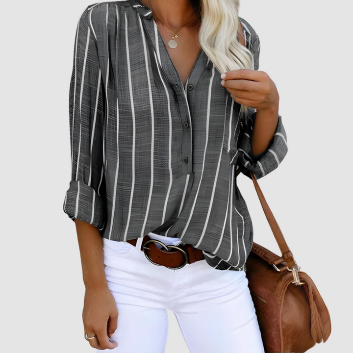 Gray and white vertical striped blouse with rolled-up sleeves, accessorized with a brown bag and white pants. Studio background.