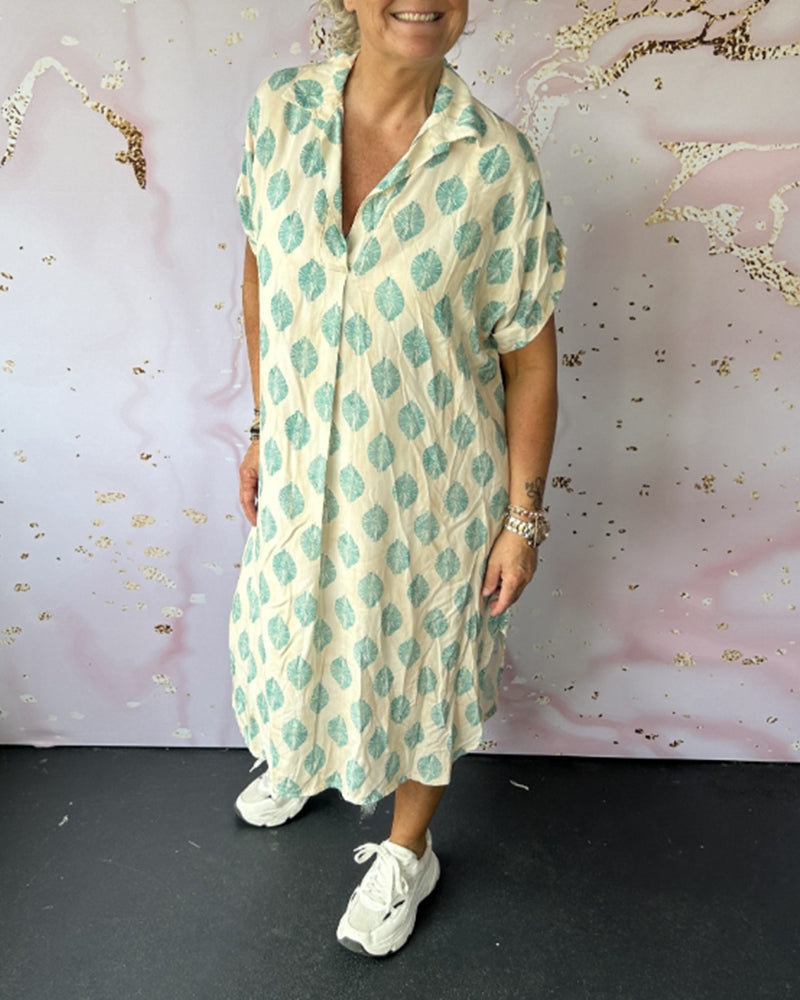 Woman wearing white and turquoise patterned dress, sneakers, in an art-decorated studio environment.