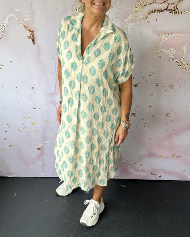Woman wearing white and turquoise patterned dress, sneakers, in an art-decorated studio environment.