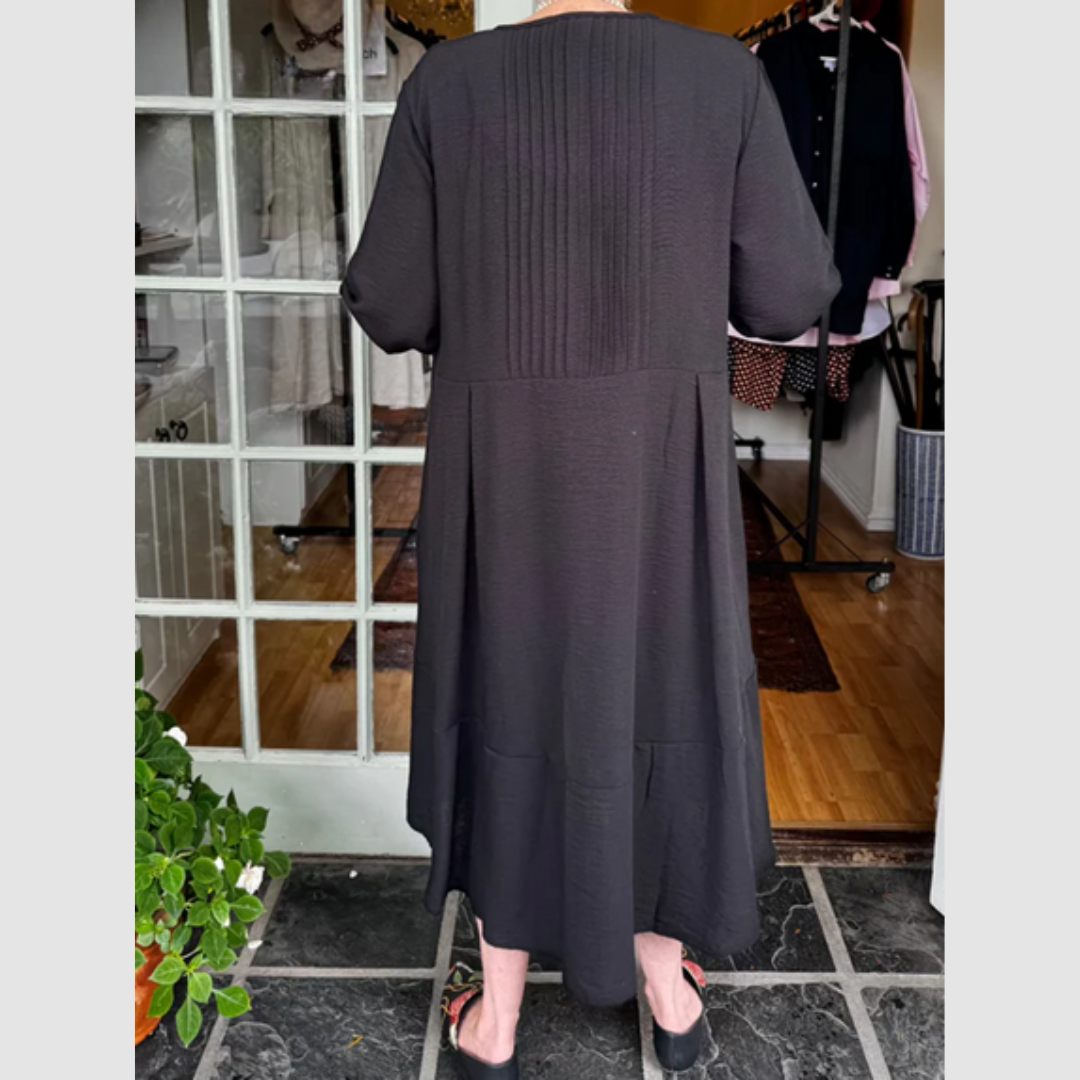 Back view of woman in a black pleated dress with irregular hem, standing near a glass door in a boutique.