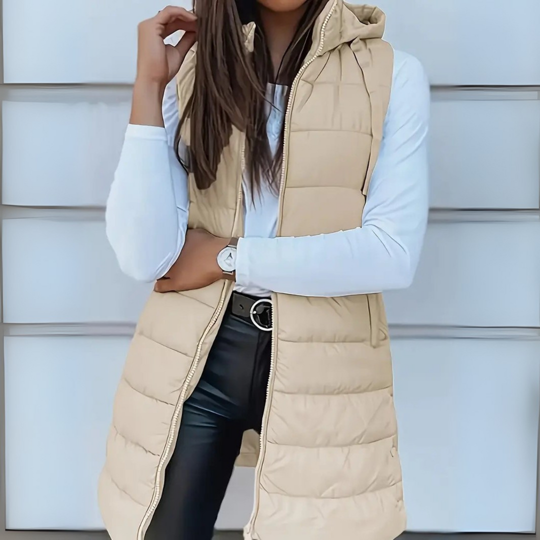 Beige sleeveless puffer vest with hood worn over white top and black pants, outdoor setting.