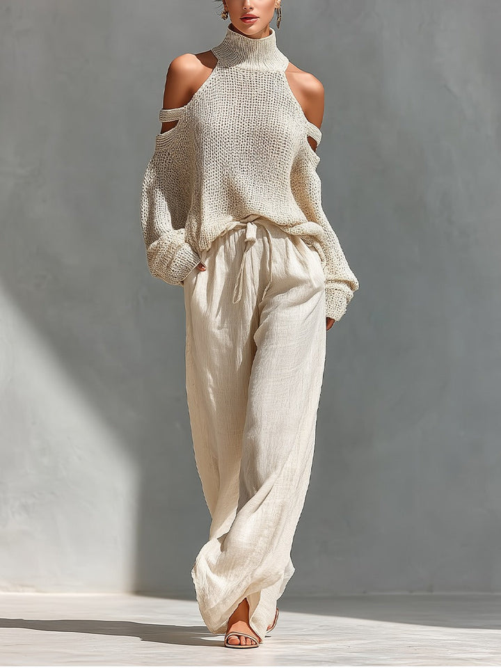 Woman wearing a cream turtleneck sweater with cold shoulder cutouts and flowing cream pants in a sunlit indoor setting.
