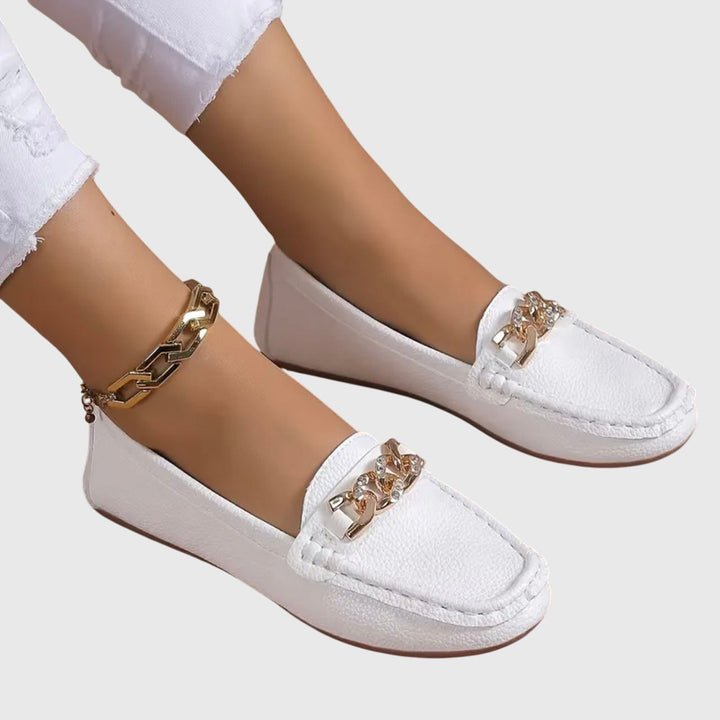 White orthopedic loafers with gold chain detail, styled with white pants on a gray background.