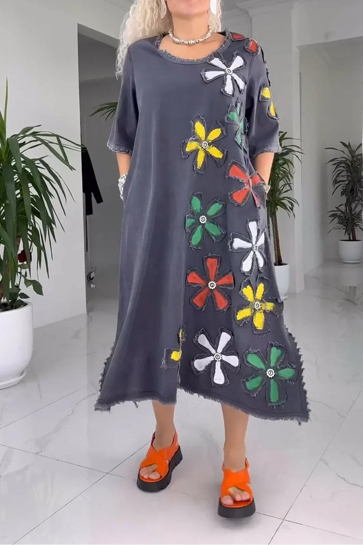 Model in dark gray floral dress with vibrant flower patches, orange sandals, in a white room with plants.