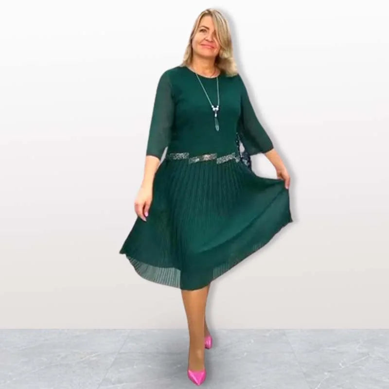 Woman wearing dark green elegant midi dress with 3/4 sleeves, pleated skirt, standing on gray tile floor in a studio.