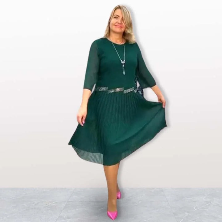 Woman wearing dark green elegant midi dress with 3/4 sleeves, pleated skirt, standing on gray tile floor in a studio.