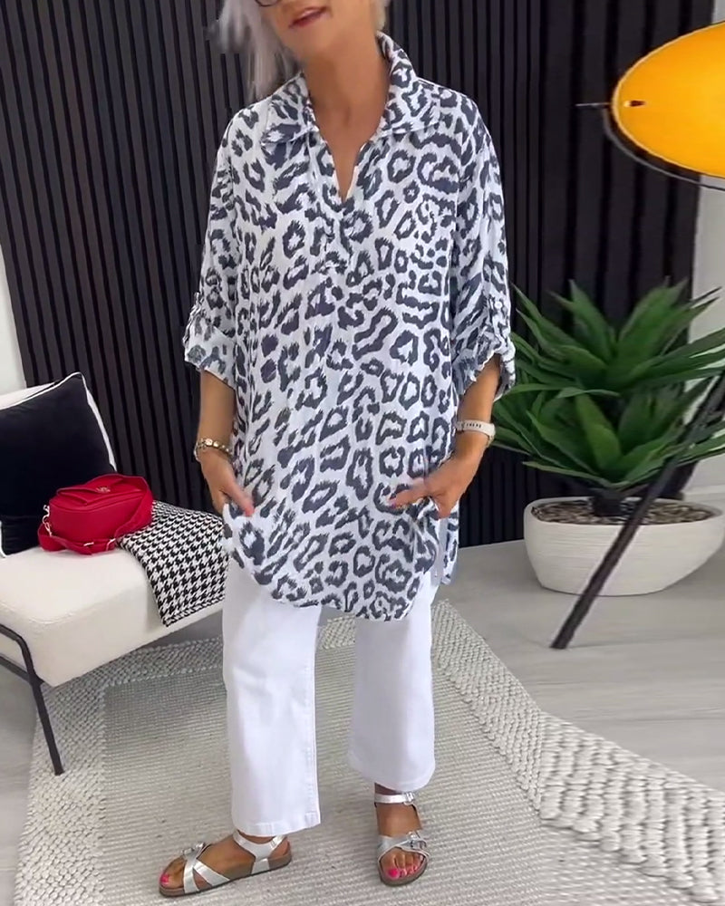 Black leopard print blouse with a collar, paired with white pants, in a contemporary indoor setting with a plant and modern decor.
