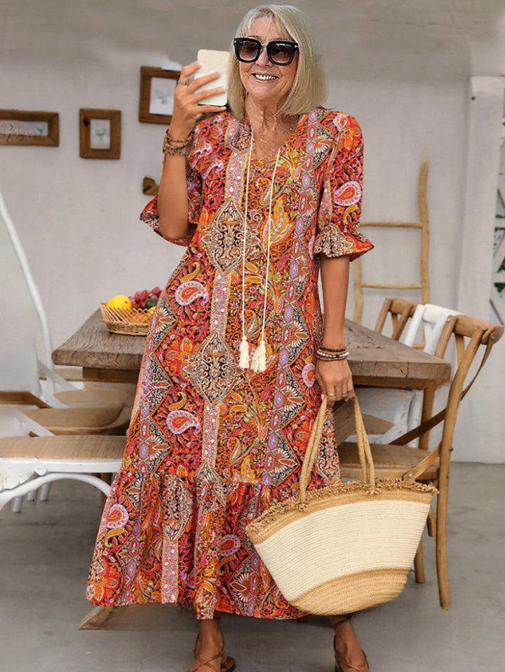 Woman in vibrant boho dress with flowy patterns, holding phone, indoor setting with wooden furniture, straw bag in hand.