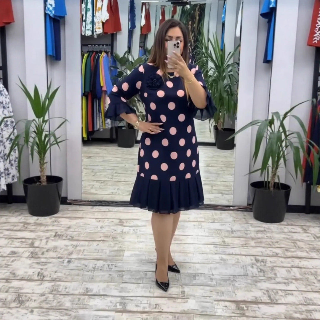 Woman taking a selfie in a boutique, wearing a navy polka dot pleated dress with pink accents, surrounded by potted plants and colorful clothing.