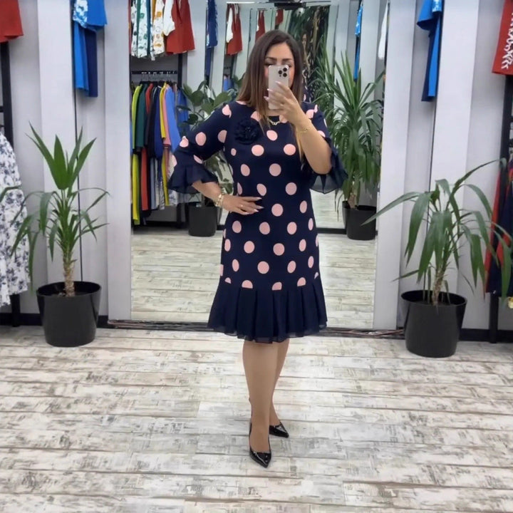 Woman taking a selfie in a boutique, wearing a navy polka dot pleated dress with pink accents, surrounded by potted plants and colorful clothing.