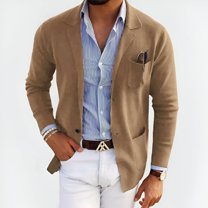 Man wearing a beige lightweight jacket over a striped shirt, paired with white pants, in a studio setting.