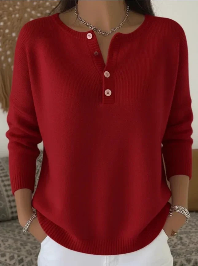 Model in red sweater with buttoned neckline, white jeans, bracelets, indoors with decorative backdrop and cushions.