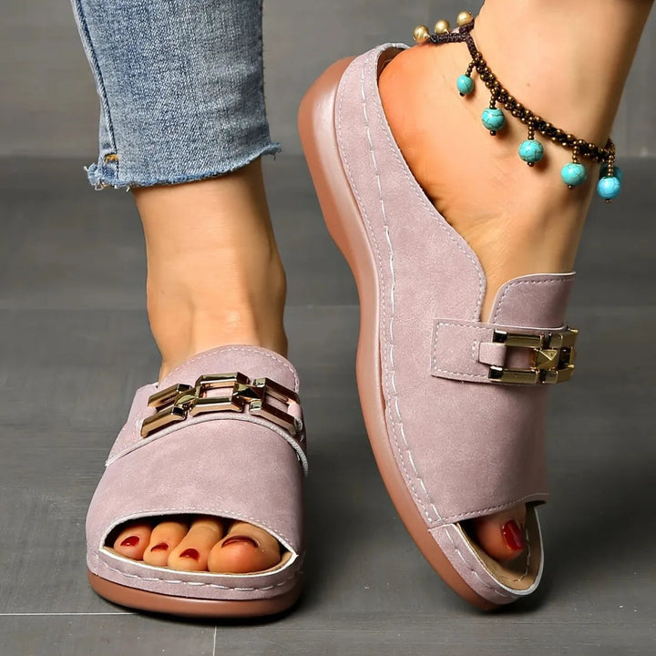 Elegant pink sandals with gold embellishments and anklet, worn indoors on grey floor.