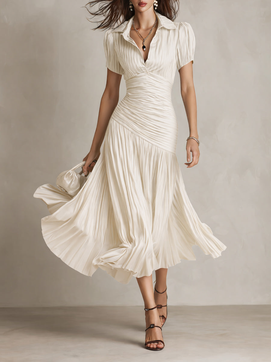 Model in flowing cream midi dress with pleated skirt and short sleeves, holding a purse in a studio setting.
