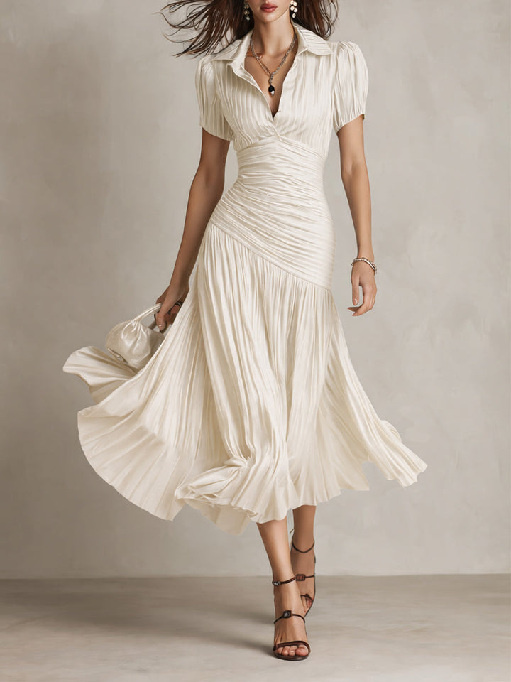 Model in flowing cream midi dress with pleated skirt and short sleeves, holding a purse in a studio setting.