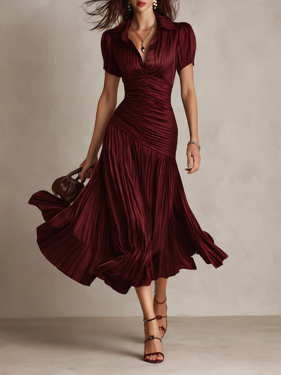Model in flowing burgundy midi dress with pleated skirt and short sleeves, holding a purse in a studio setting.