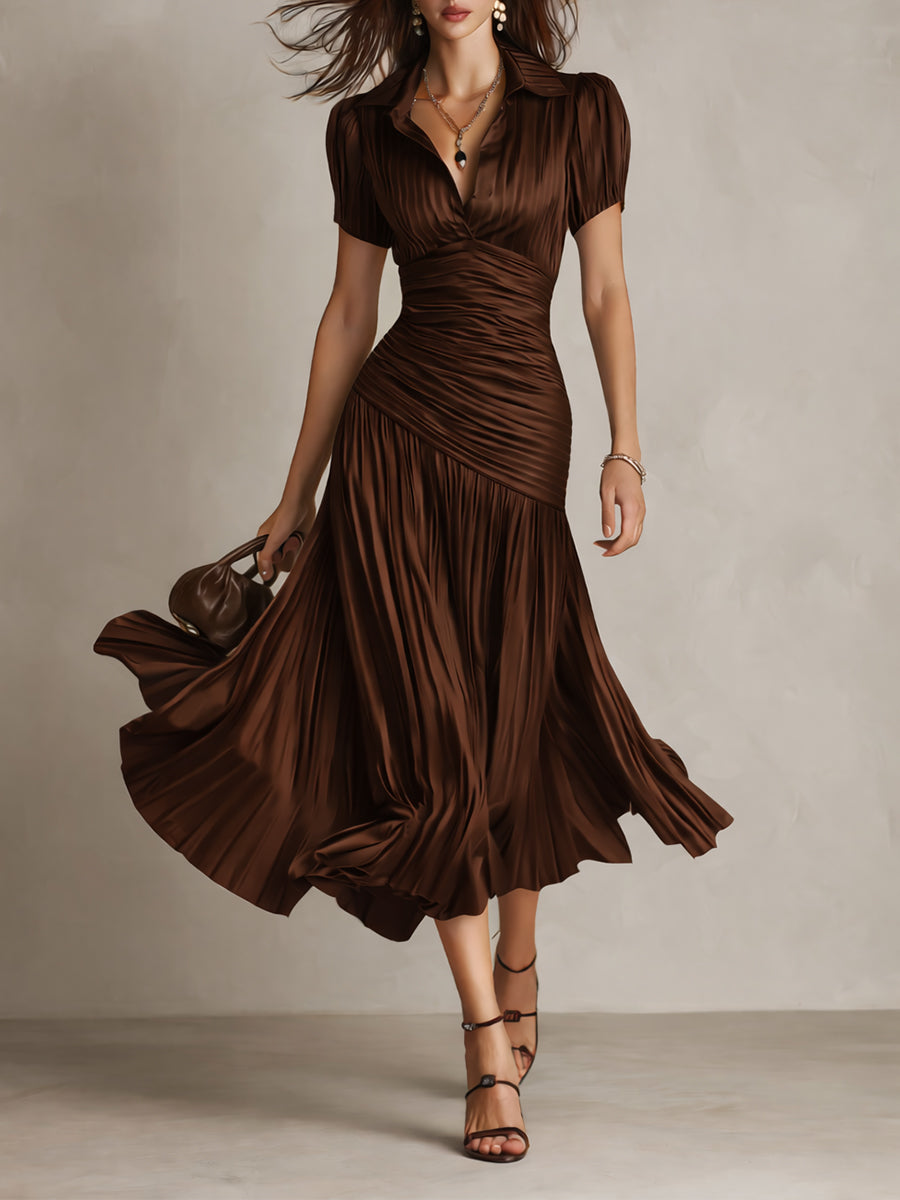 Model in flowing brown midi dress with pleated skirt and short sleeves, holding a purse in a studio setting.