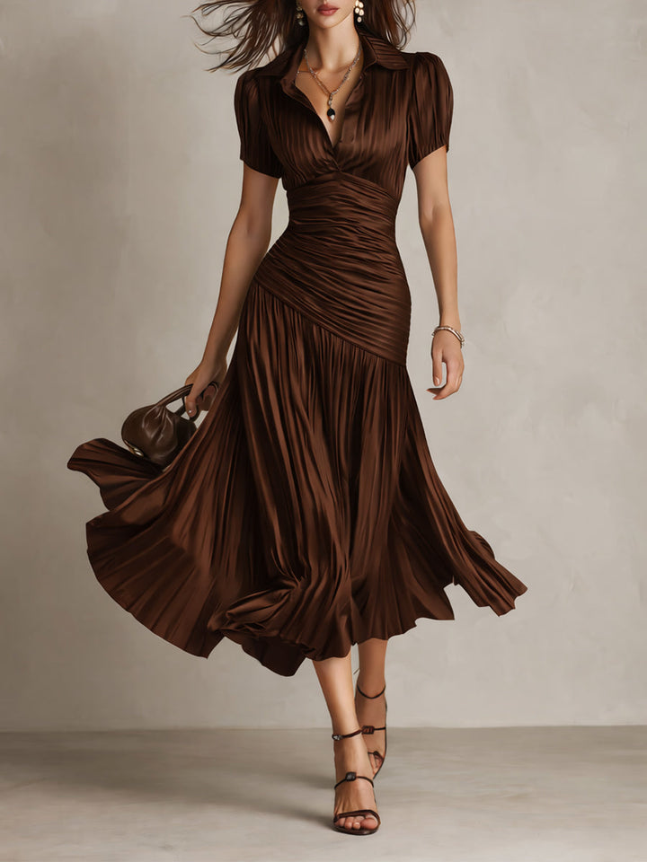Model in flowing brown midi dress with pleated skirt and short sleeves, holding a purse in a studio setting.