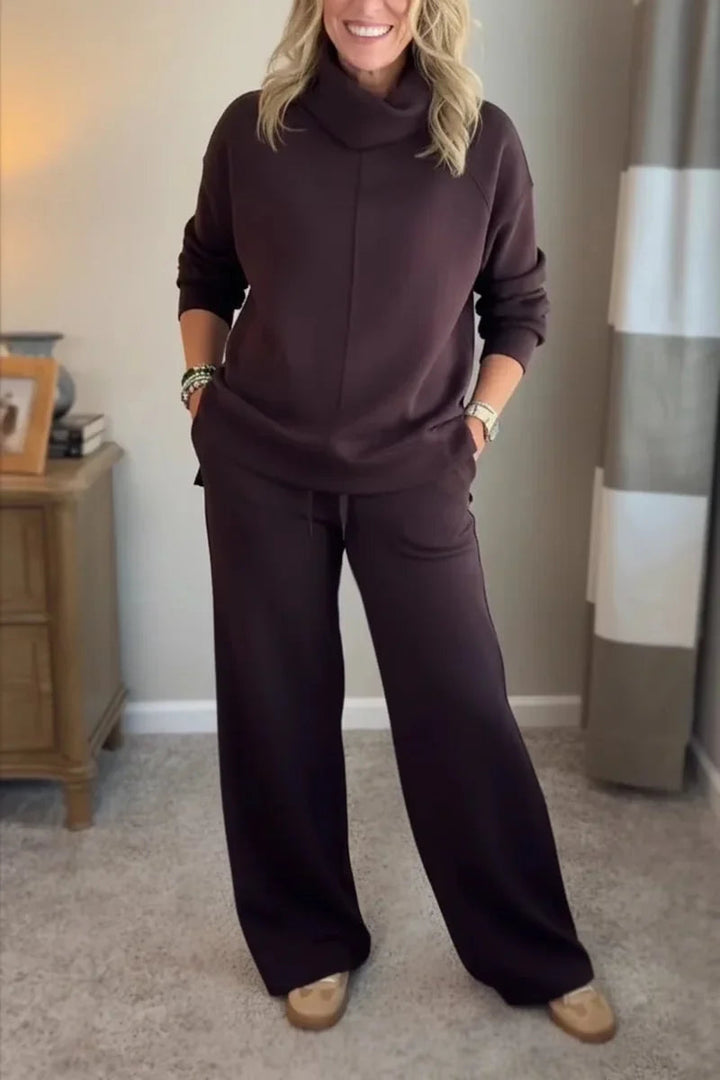 Woman wearing a dark brown comfy set with long sleeves and wide-leg pants, standing in a carpeted room near a wooden table.