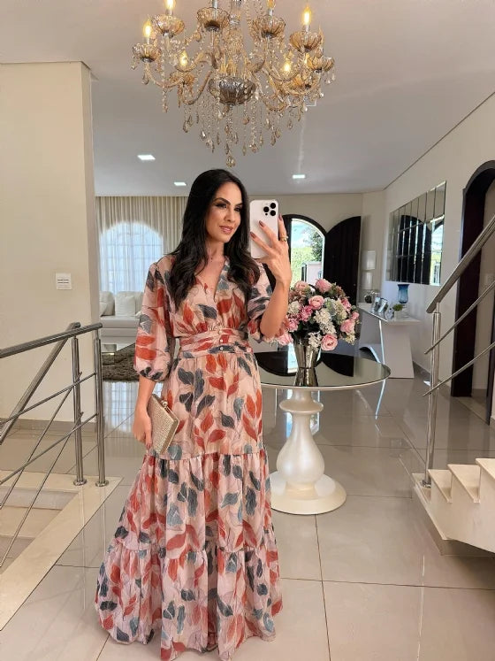 Model in a flowing floral maxi dress with autumn tones, holding a clutch in an elegant living room under a chandelier.