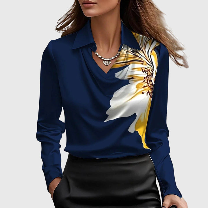 Navy blue blouse featuring a large yellow and white flower print, draped neckline, and long sleeves, with a black skirt on neutral backdrop.