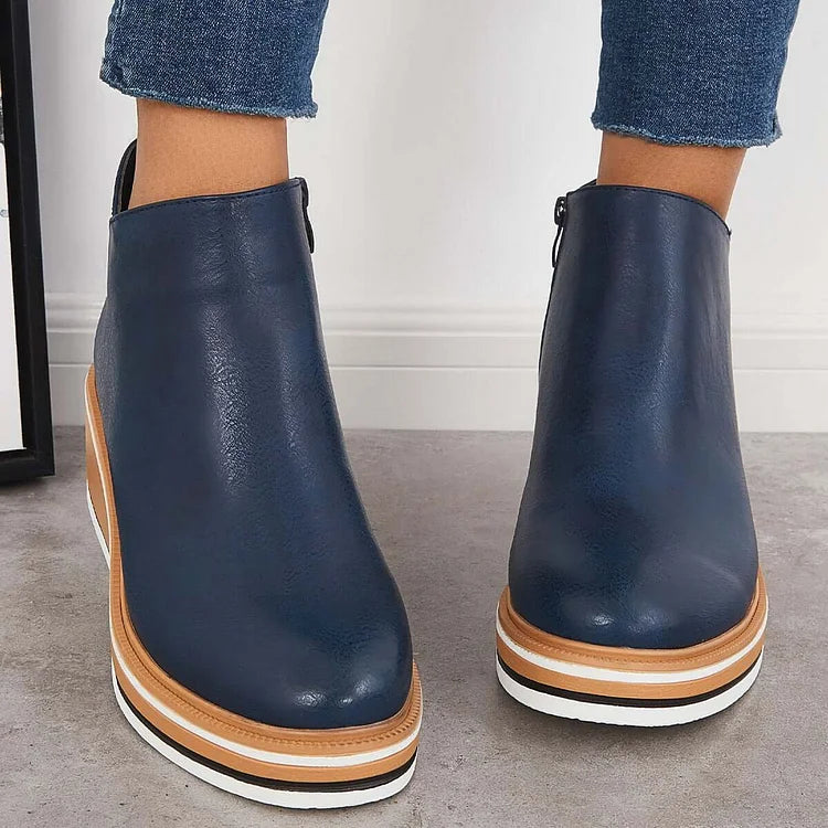 Front view of navy blue luxe boots with wedge soles, worn with frayed jeans, on a gray floor.