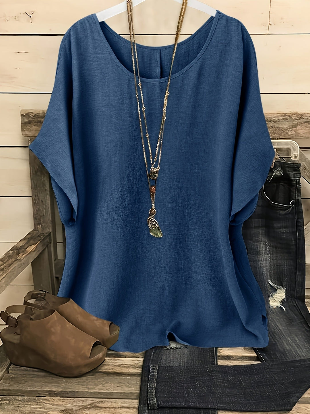 Navy blue loose summer top on hanger with necklace, sandals, and jeans on rustic bench.