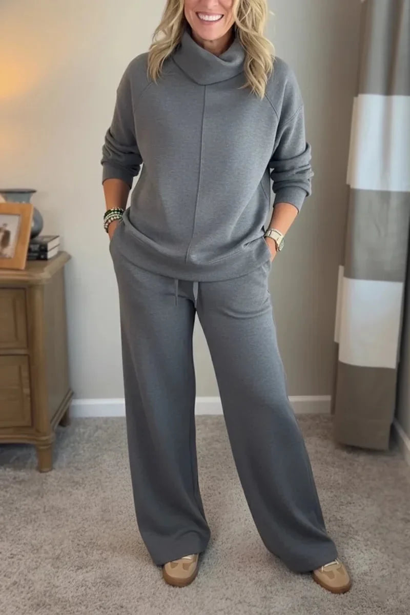 Woman wearing a gray comfortable set with long sleeves and wide-leg pants, standing in a carpeted room near a wooden table.