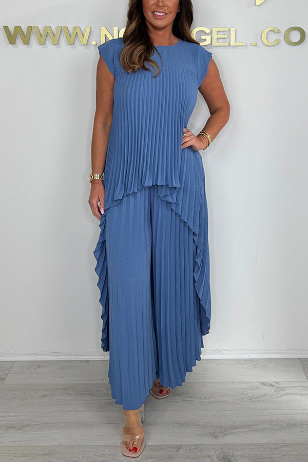 Woman wearing a blue pleated midi top and wide-leg pants set, standing in a studio with a neutral wall and floor.