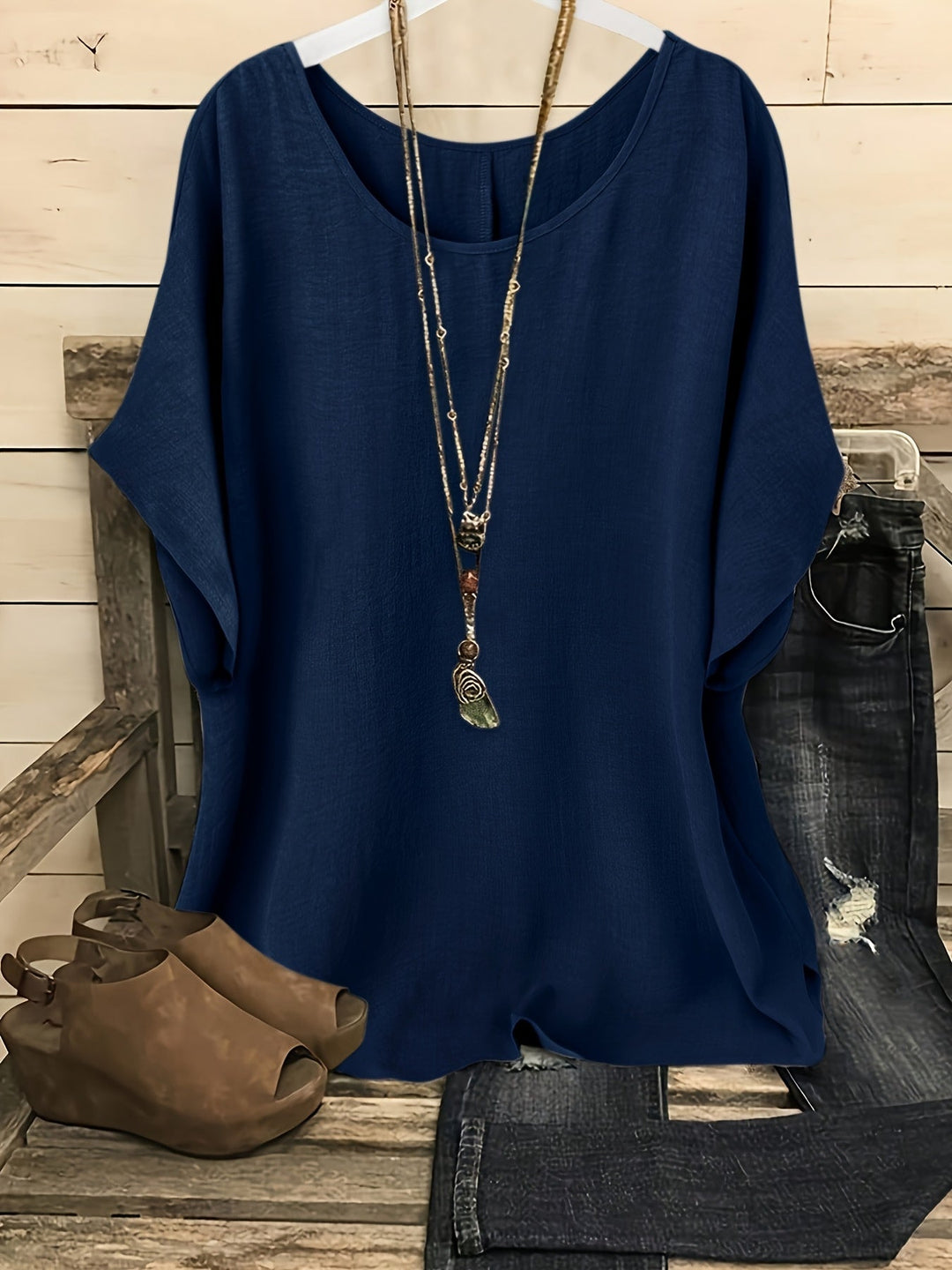 Indigo blue loose summer top on hanger with necklace, sandals, and jeans on rustic bench.