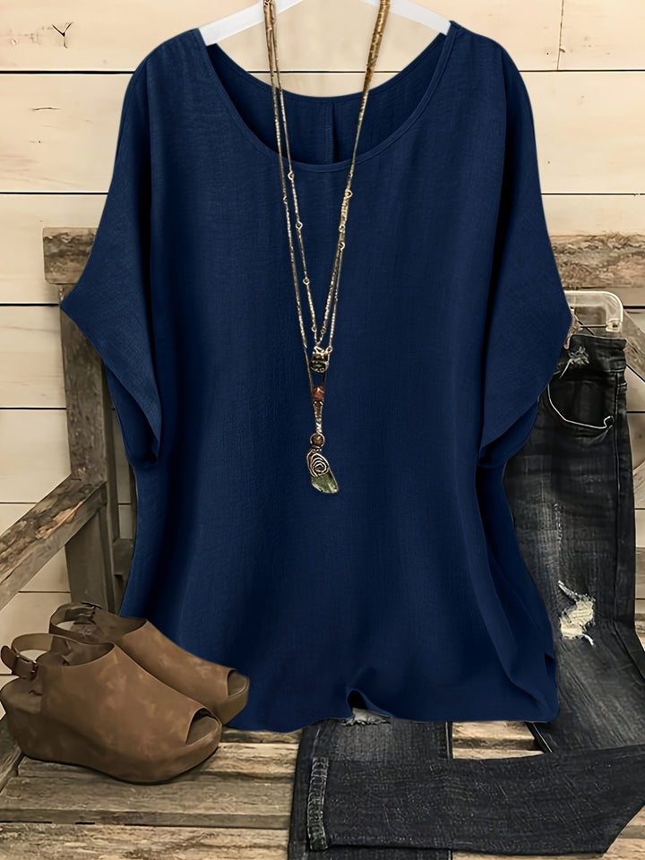 Indigo blue loose summer top on hanger with necklace, sandals, and jeans on rustic bench.