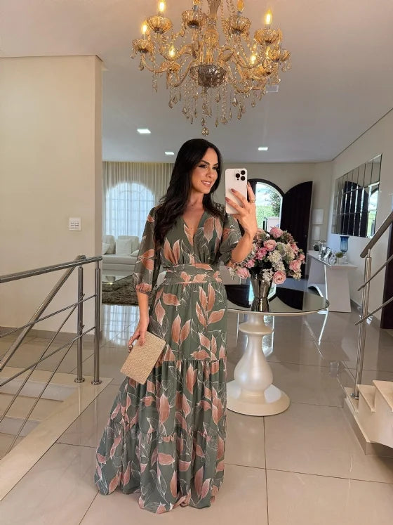 Model in a green and pink floral maxi dress, clutch in hand, in a stylish living room with a chandelier.