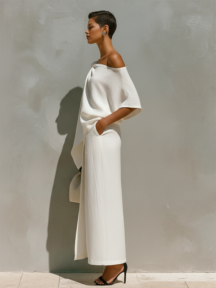 Side profile of a model in a chic white off-shoulder dress, hands in pockets, against a textured grey backdrop.