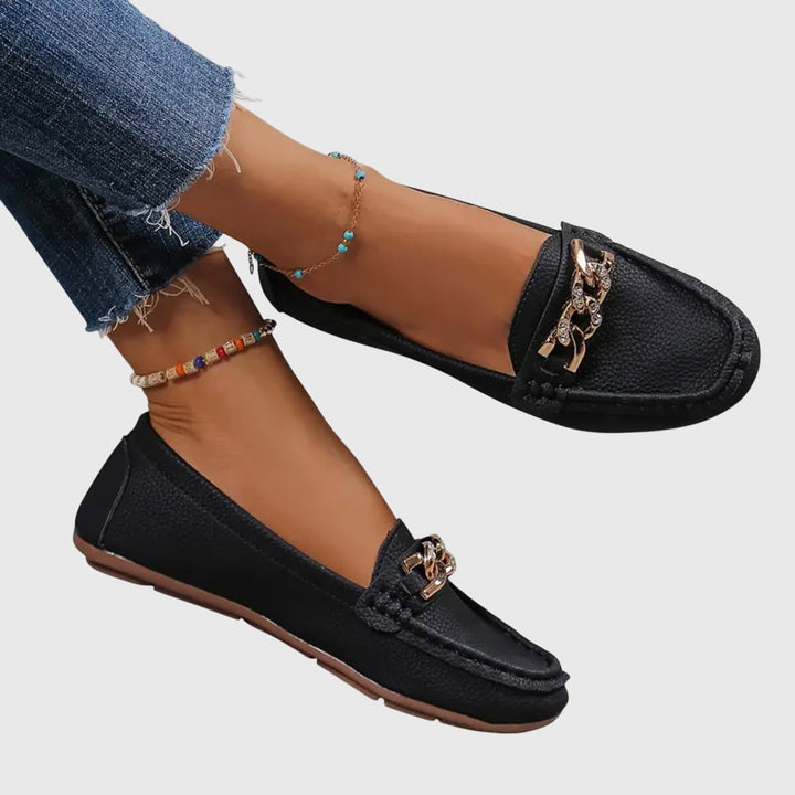 Black orthopedic loafers with gold chain detail, paired with denim jeans, on a gray background.