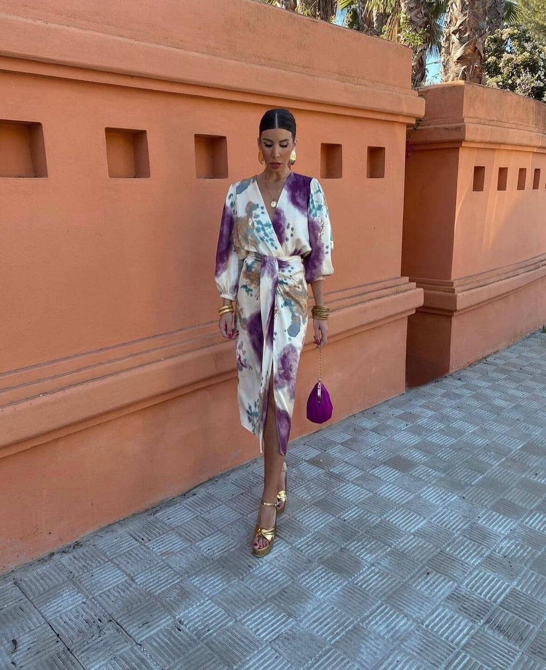 Flowing dress with purple and blue tones, worn by a woman on a tiled pathway next to an orange wall with cutouts.