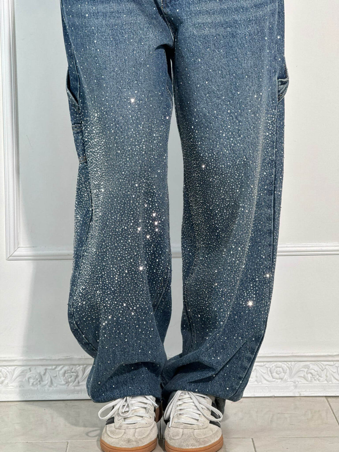 Close-up of dark blue trousers showcasing diamond embellishments and white sneakers in a bright studio.