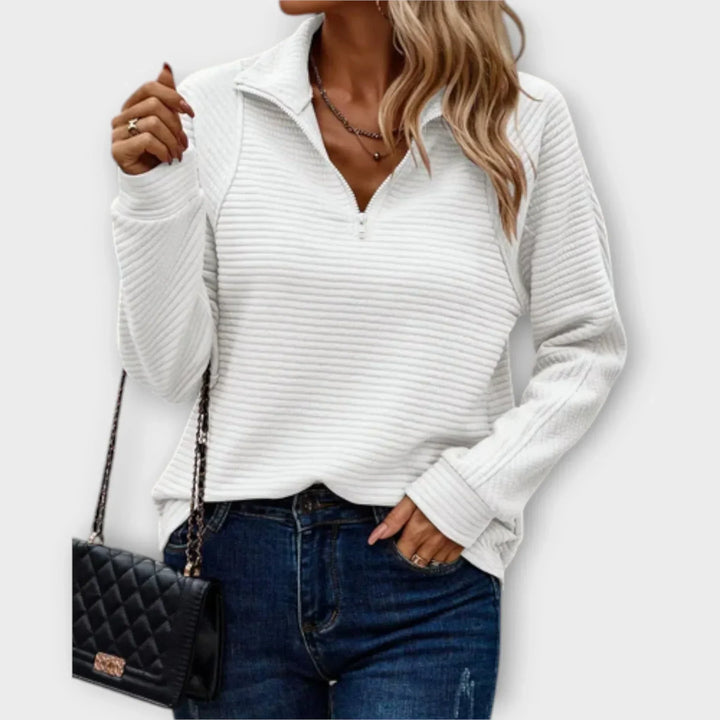 Woman in a white half-zip pullover with ribbed texture, holding a black quilted purse, wearing jeans, on a plain backdrop.