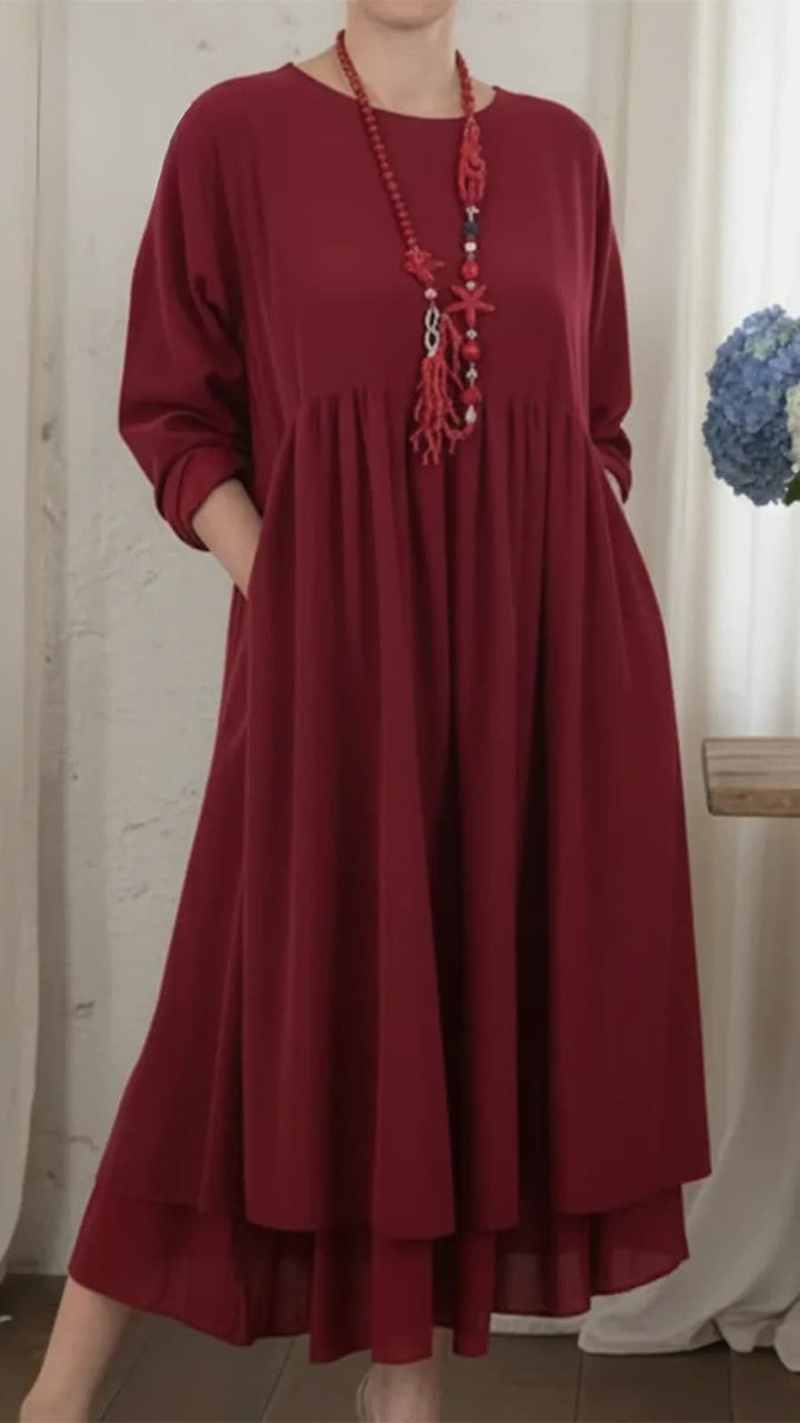 Burgundy maxi dress with long sleeves and a relaxed cut, adorned with a red beaded necklace, placed in an indoor setting with soft lighting.