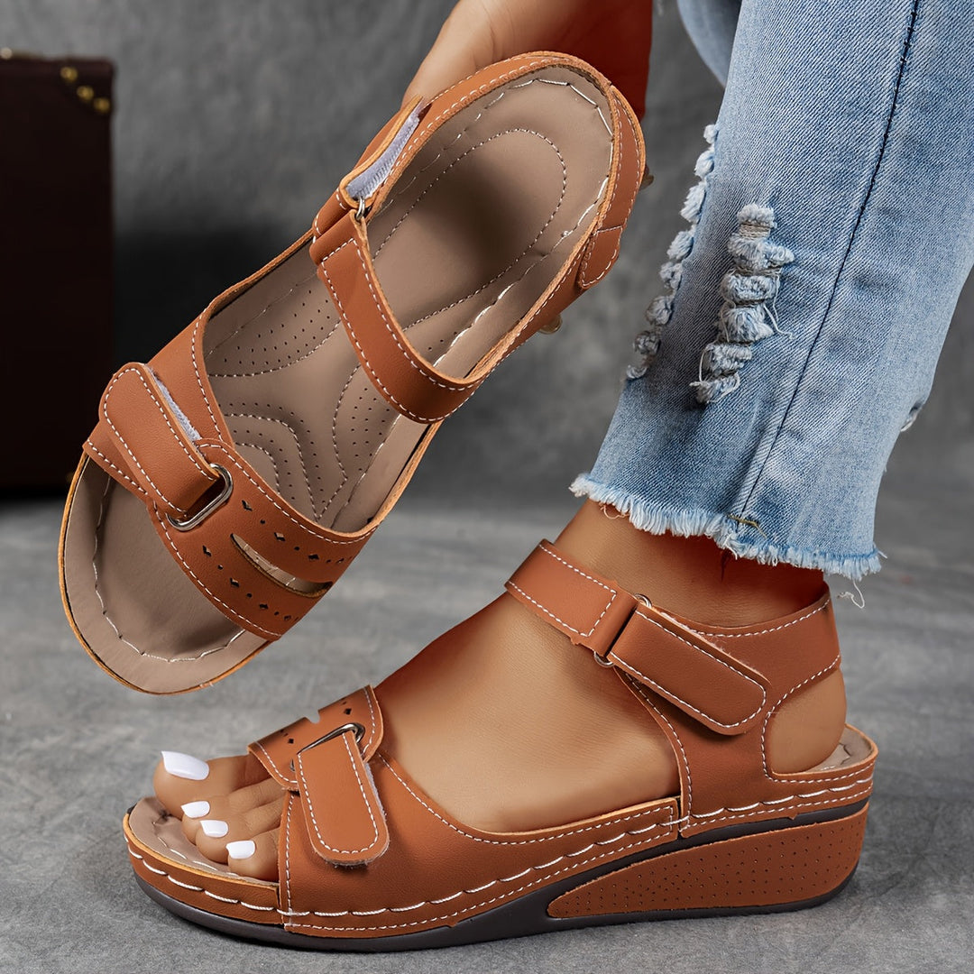Brown orthopedic sandal featuring comfortable straps and soft footbed, paired with ripped jeans on a gray stone floor.