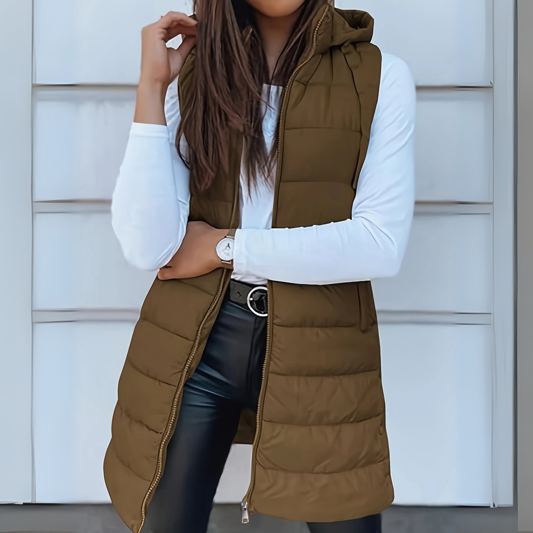 Olive green sleeveless puffer vest with hood worn over white top and black pants, outdoor setting.